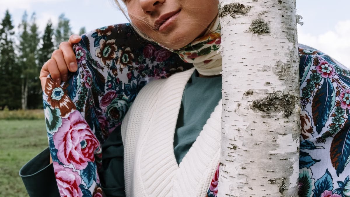 A woman in a colorful floral scarf stands beside a birch tree in a lush outdoor setting, exuding warmth and style.